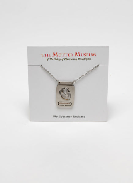 A white card with the Mütter Museum logo on it and "Wet Specimen Necklace" written on the bottom. There is a stainless steel chain with a pendant that depicts a jar with a laser-etched heart inside it. There is a label on the jar that reads Mütter Museum.