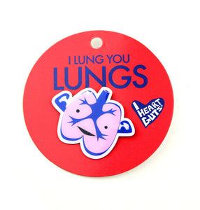 A red card with "I Lung You Lungs" written on it in blue letters. There is a pink and blue enamel pin in the shape of lungs with two arms depicting muscles. There are two eyes and a smile.