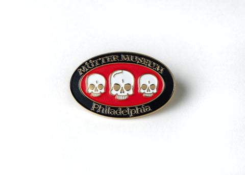 An enamel pin in the shape of an oval with three skulls in the center. The outer rim is black with "Mütter Museum, Philadelphia" written in it and the skulls are white with a red background.