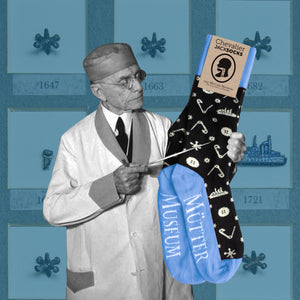 A pair of black socks with blue heels, toes, and ankles. There are white pictures of safety pins, buttons, and a ship on it. There is a photo of Dr. Jackson pointing at the socks with an image of one of the Chevalier Jackson drawers in the background.