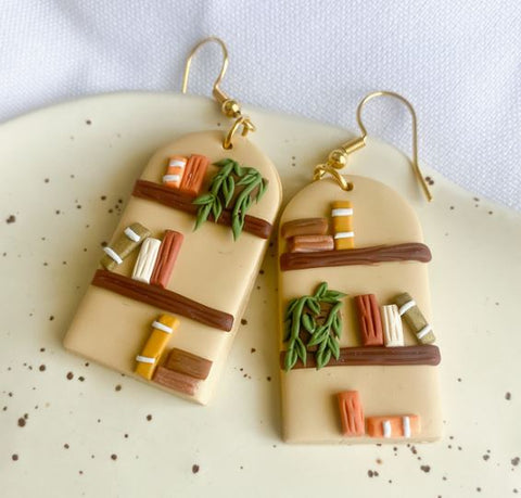 A pair of arched being clay earrings with gold ear wires. The beige arches have bookshelves with colorful books and plants on them, all made out of clay.