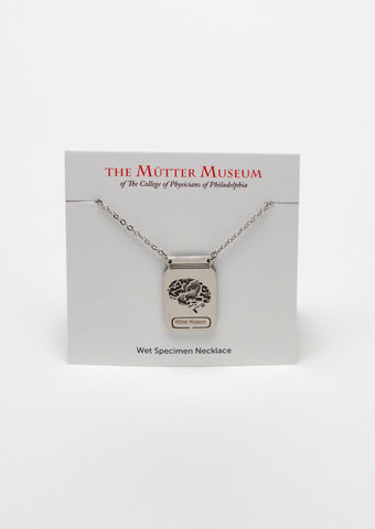 A white card with the Mütter Museum logo on it and "Wet Specimen Necklace" written on the bottom. There is a stainless steel chain with a pendant that depicts a jar with a laser-etched brain inside it. There is a label on the jar that reads Mütter Museum.