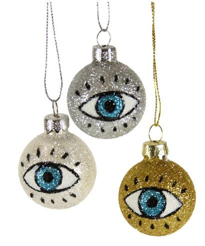 Three round ornaments; one sparkly silver, sparkly white, and sparkly gold. Each one depicts an eyeball with lashes and blue irises.