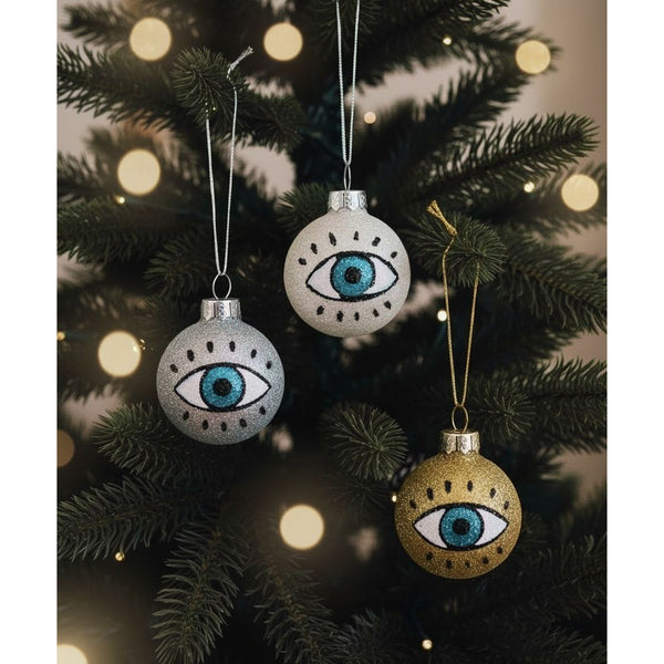 Three round ornaments; one sparkly silver, sparkly white, and sparkly gold. Each one depicts an eyeball with lashes and blue irises.