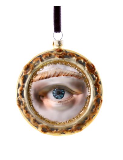 A glass ornament in the shape of a flat circle with a brown frame. There is gold glitter on the inside of the frame, with a depiction of an eyeball and eyebrow. The iris is blue and the skin is pale with bronze highlights.