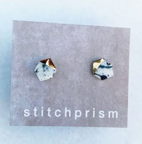 A pair of hexagon shaped stud earrings made out of white clay with black splatters and one edge dipped in gold.