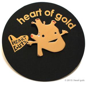 A black card with "Heart of Gold" written on it in gold. There is an enamel pin in the shape of an anatomical heart in gold with two eyes and a smile.