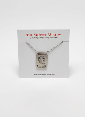 A white card with the Mütter Museum logo on it and "Wet Specimen Necklace" written on the bottom. There is a stainless steel chain with a pendant that depicts a jar with a laser-etched heart inside it. There is a label on the jar that reads Mütter Museum.