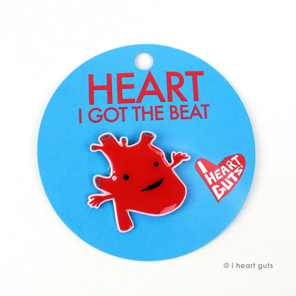A blue card with "Heart I Got the Beat" in red letters written on it. There is a red enamel pin in the shape of an anatomical heart with two eyes and a smile.