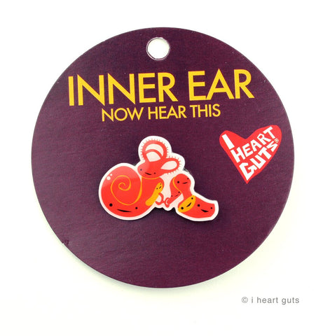 A maroon card with 'Inner Ear Now Hear This" written in yellow letters. There is an enamel pin depicting the cochlea in red and yellow. Each part has its own face on it consisting of two eyes and a smile.