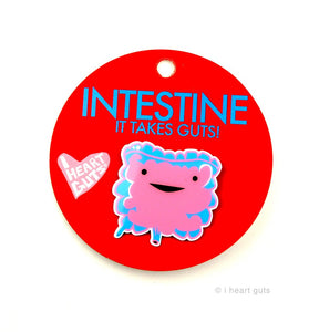 A red card with "Intestine It Takes Guts" written in blue letters. There is a blue and pink enamel pin in the shape of an intestine with two eyes and a smile.