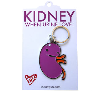 A white card with "Kidney When Urine Love" written on it in purple letters. There is a purple and orange enamel keychain in the shape of a kidney with a smile and two eyes on it.