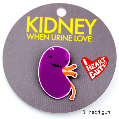 A grey card with "Kidney When Urine Love" in yellow letters written on it. There is a purple and orange enamel pin in the shape of a kidney on it.