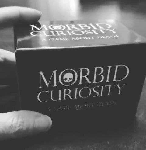 A black and white photo of a hand holding a boxed game. The box has "Morbid Curiosity: A Game About Death" written on it in white and there is an illustration of a white skull in the O of Morbid.