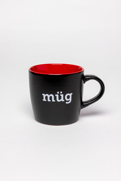 A glossy black mug with a handle and a red inside. On the front of the mug is "müg" in white and on the back is the Mütter Museum logo.