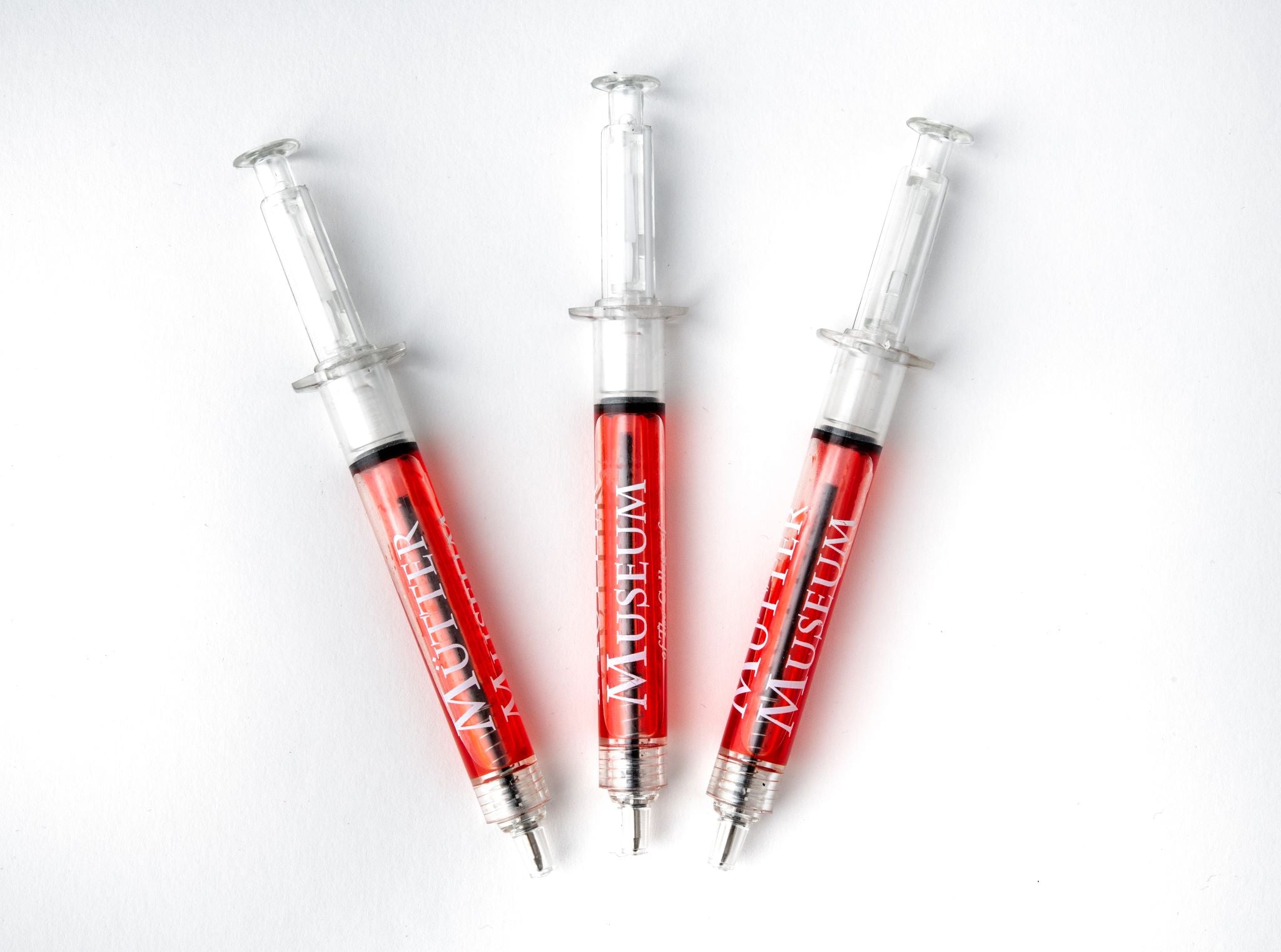 Three plastic pens with a click top in the shape of a syringe. There is a red liquid inside the pen and the Mütter Museum logo is on the barrel.