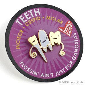 A card with a black rim and purple background that says "Teeth Flossin' Ain't Just for Gangstas" and a label for incisor, cuspid, and molar. There is a white and gold enamel pin of all three teeth next to each other, each with eyes and a smile.